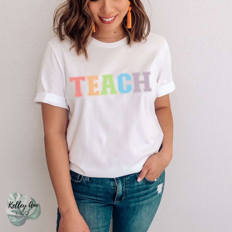 May include: White t-shirt with the word "TEACH" in rainbow colors. The letters are outlined in white.