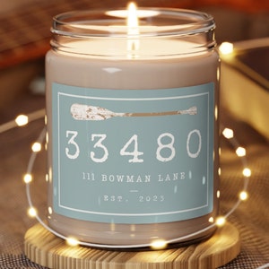 May include: A brown candle in a clear glass jar with a blue label. The label has a white oar and the text "33480 111 BOWMAN LANE EST. 2023".