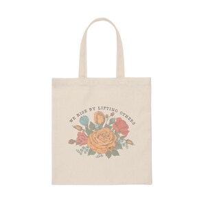 May include: A beige tote bag with a floral design and the text "We rise by lifting others".