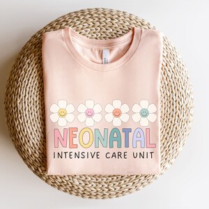 May include: A pink t-shirt with a rainbow colored word "NEONATAL" and the words "INTENSIVE CARE UNIT" in black. The word "NEONATAL" is surrounded by five white daisies with yellow centers and smiling faces.
