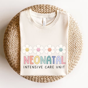 May include: A cream-colored t-shirt with a row of five colorful daisy flowers with smiley faces. The text "NEONATAL INTENSIVE CARE UNIT" is written in rainbow colors below the flowers.