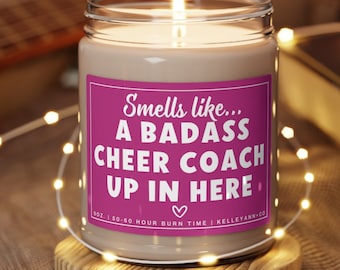 May include: A brown glass candle with a pink label that reads "Smells like... A BADASS CHEER COACH UP IN HERE" with a heart symbol. The candle is on a wooden coaster.