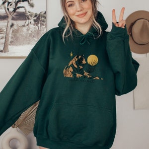 May include: A dark green hooded sweatshirt with a graphic print of a tiger sitting in a desert landscape with a yellow moon and stars. The text "wild spirit" is printed below the tiger.
