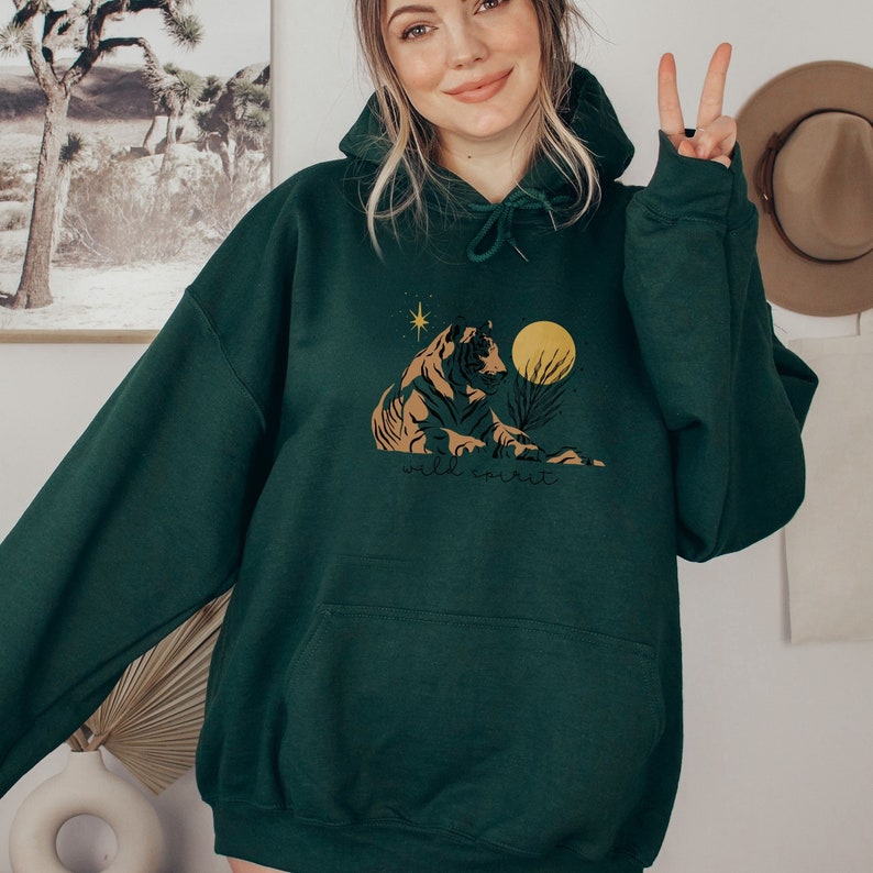 May include: A dark green hooded sweatshirt with a graphic print of a tiger sitting in a desert landscape with a yellow moon and stars. The text "wild spirit" is printed below the tiger.
