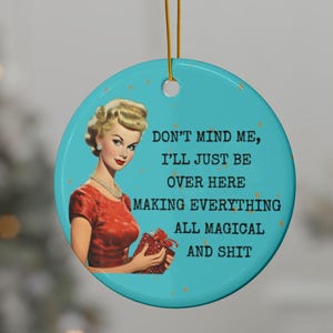 Könnte beinhalten: Ein rundes, türkisfarbenes Keramikornament mit einer Vintage-Illustration einer blonden Person in einem roten Kleid, die ein Geschenk hält. Der Text lautet: DON'T MIND ME, I'LL JUST BE OVER HERE MAKING EVERYTHING ALL MAGICAL AND SHIT. Mit goldener Schnur.