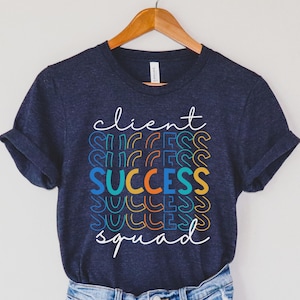 May include: Navy blue t-shirt with the words "Client Success Squad" in white script. The word "Success" is stacked in colorful, retro-style lettering. The shirt is hanging on a wooden hanger.
