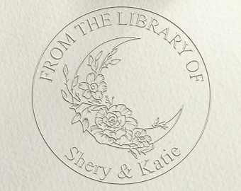 May include: A silver self-inking stamp with a round design featuring a crescent moon with floral accents. The text "From the Library of Shery & Katie" is printed around the moon.