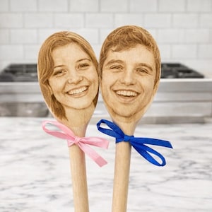 Puede incluir: Dos cucharas de madera con caras grabadas, una con una cinta rosa y otra con una cinta azul. Las cucharas presentan caras sonrientes y probablemente son utensilios de cocina personalizados.
