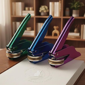 Puede incluir: Tres sellos metálicos de colores verde, azul y morado descansan sobre un libro blanco con texto en relieve. Los sellos tienen un diseño de palanca y bases circulares doradas. El libro está sobre una superficie de cuero marrón.