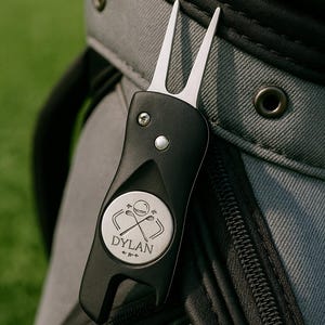 Pode incluir: Uma ferramenta preta para reparar divots de golfe com um garfo prateado e um emblema circular. O emblema apresenta tacos de golfe cruzados e o nome "DYLAN". A ferramenta está presa a um saco de golfe cinza, com fundo verde.