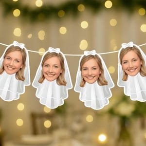 Puede incluir: Cuatro adornos blancos recortados con forma de cabeza de novia colgados de una cuerda. Cada recorte presenta a una mujer sonriente con cabello rubio, un velo blanco y un lazo. El fondo está borroso con luces bokeh, sugiriendo un ambiente festivo.