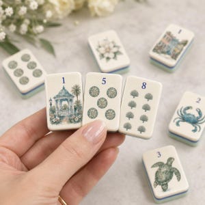 Puede incluir: Un conjunto de fichas de mahjong rectangulares con diversas ilustraciones, incluyendo un cenador, un cangrejo, una tortuga marina, una flor y valores numéricos. Las fichas son de color blanco roto con diseños en tinta azul y verde.