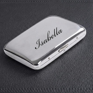 May include: A rectangular silver-toned metal case with rounded corners and a polished finish. The name Isabella is engraved in a black script font across the center of the top surface. A small metal latch is visible on the side of the case.