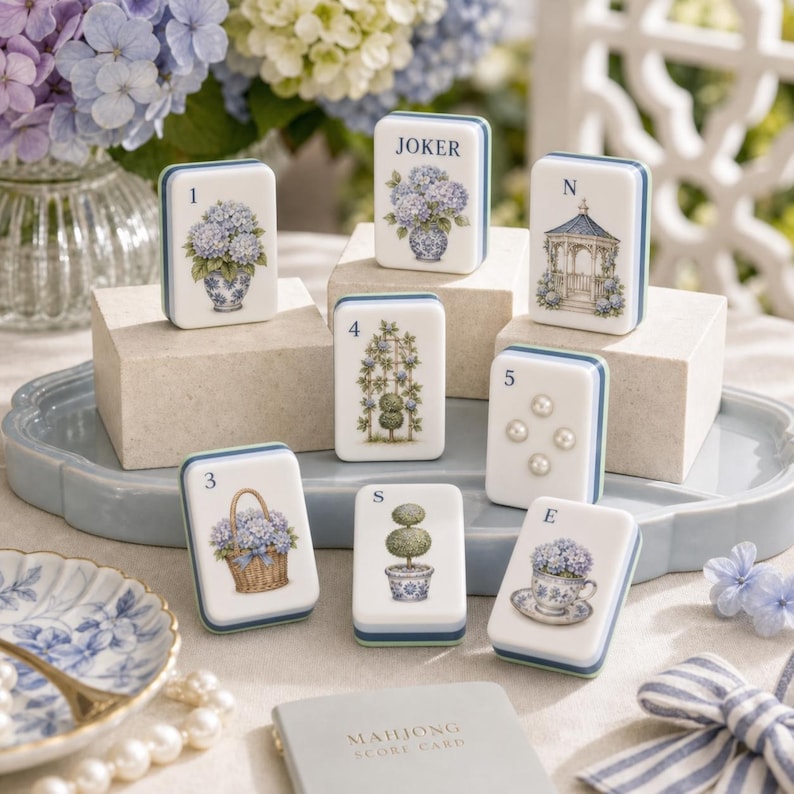 Puede incluir: Un conjunto de fichas de mahjong rectangulares con ilustraciones florales en azul y blanco, que incluyen hortensias en jarrones, un cenador, un topiario y una taza de t&eacute;. Cada ficha muestra una letra o n&uacute;mero, con cuatro perlas blancas.
