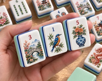 Op de afbeelding: Drie rechthoekige mahjongtegels met witte voorzijden en lichtgroene en blauwe zijkanten. De tegels bevatten kleurrijke illustraties van een brug, een ijsvogel op een bamboetak en een bloemenvaas, elk gemarkeerd met een rood of blauw cijfer.