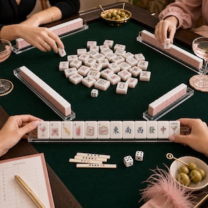 K&ouml;nnte beinhalten: Ein Mahjong-Spielset mit wei&szlig;en Spielsteinen, die rosa und schwarze Illustrationen wie Zahlen, Cocktail-Grafiken und chinesische Schriftzeichen zeigen. Das Set enth&auml;lt transparente Acryl-Halterungen, zwei wei&szlig;e W&uuml;rfel und einen Punkteblock.