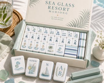 May include: A Sea Glass Resort Mahjong game set in a cream-coloured box with palm leaf illustrations. The set includes white tiles featuring blue numbers, Chinese characters, and coastal icons like sailboats and flowers, plus blue tile racks.