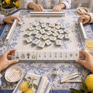 May include: A Mahjong set featuring white tiles with blue and yellow accents, including illustrations of a sailboat, house, tree, and sun, alongside Chinese characters. The set includes a tile rack, two white dice, and scoring sticks on a blue and white patterned surface.