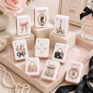 Puede incluir: Una colección de nueve fichas rectangulares blancas con diversas ilustraciones que incluyen un lazo rosa, una silueta, la Torre Eiffel, flores, perlas, un bolso negro, un bolso acolchado rosa, una escena de café y una flor de lis con el texto JOKER.