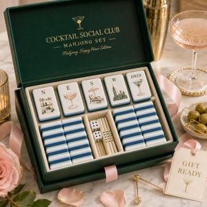 K&ouml;nnte beinhalten: Ein dunkelgr&uuml;ner Mahjong-Set-Kasten mit goldenem Schriftzug COCKTAIL SOCIAL CLUB MAHJONG SET. Im Inneren befinden sich wei&szlig;e Spielsteine mit Abbildungen eines Barwagens, Cocktails, Liegestuhls, einer Skyline und eines Martinis sowie blaue Streifensteine.
