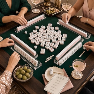 K&ouml;nnte beinhalten: Ein Mahjong-Spielset mit blassrosa Spielsteinen mit detaillierten Illustrationen, angeordnet auf einer dunkelgr&uuml;nen Filzoberfl&auml;che. Das Set enth&auml;lt transparente Acryl-Spielsteinhalter, zwei wei&szlig;e W&uuml;rfel und eine Cocktail-Men&uuml;karte.