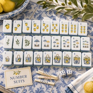 May include: A mahjong tile set featuring three rows of nine white tiles with blue and green illustrations of lemons, beach umbrellas, and floral tote bags. The set includes a card labeled NUMBER SUITS, three small counting sticks, and two dice.