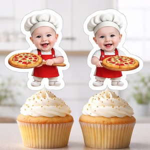 May include: Two cupcake toppers featuring a smiling baby chef holding a pizza on a wooden paddle. The baby wears a white chef's hat, a red apron, and a white shirt. Two cupcakes with white frosting and gold sprinkles are below.
