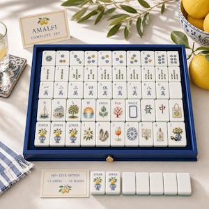 May include: A mahjong set in a blue wooden tray featuring white tiles with custom illustrations of lemons, coastal scenes, and traditional symbols. The set includes a card labeled Amalfi Complete Set and a 160-tile option card with extra blank tiles.