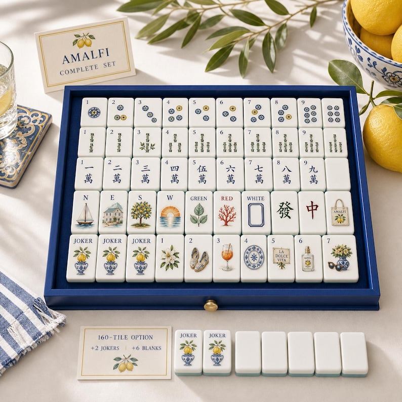 May include: A mahjong set in a blue wooden tray featuring white tiles with custom illustrations of lemons, coastal scenes, and traditional symbols. The set includes a card labeled Amalfi Complete Set and a 160-tile option card with extra blank tiles.