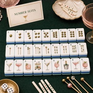 K&ouml;nnte beinhalten: Ein Set aus rechteckigen Mahjong-Spielsteinen, angeordnet in drei Reihen mit den Zahlen eins bis neun. Die Steine zeigen goldfarbene Punkte, vertikale Linien und bunte Cocktail-Illustrationen. Eine Karte mit der Aufschrift NUMBER SUITS liegt bei.