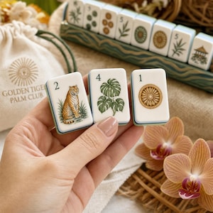 May include: Three white rectangular mahjong tiles featuring illustrations of a tiger, a monstera plant, and a circular emblem, each marked with a number. The tiles are held in front of a drawstring bag labeled GOLDEN TIGER PALM CLUB.