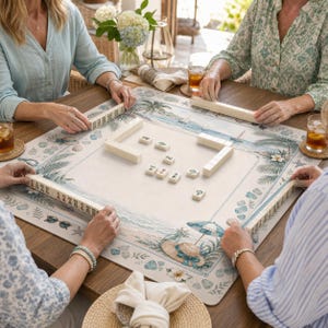 Op de afbeelding: Een vierkante mahjongmat met een kuststrandtafereel met palmbomen, een zonnehoed en een parasol. De mat ligt op een houten tafel met ivoorkleurige mahjongtegels die in rijen zijn gerangschikt en verspreid in het midden liggen voor een spel.