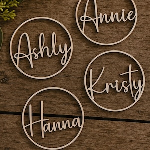 Peut inclure: Quatre étiquettes de nom en bois avec les noms "Ashly", "Annie", "Kristy" et "Hanna" en écriture cursive. Chaque nom est à l'intérieur d'un cadre circulaire. Les étiquettes sont disposées sur une surface en bois foncé.