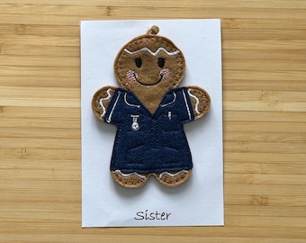 May include: A gingerbread person ornament wearing a blue uniform with the word "Sister" embroidered on it.