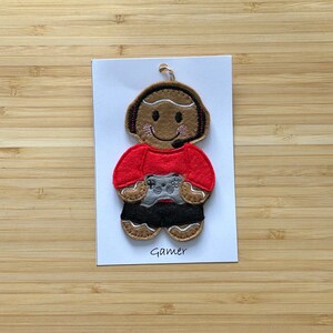 May include: A felt gingerbread person ornament with brown skin, red shirt, black trousers, and a grey video game controller. The ornament has a headset and a smiling face. The text "Gamer" is printed below the ornament.