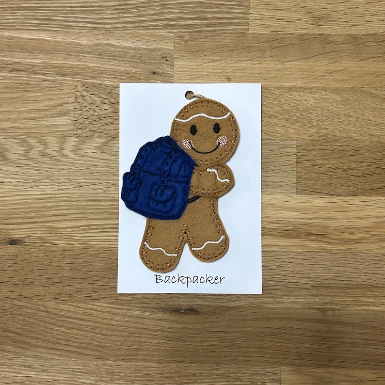 May include: A gingerbread person with a blue backpack, embroidered on felt. The gingerbread person is smiling and has white icing details. The text "Backpacker" is below the gingerbread person.