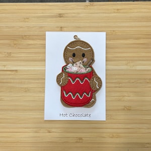 May include: A gingerbread man holding a red mug of hot chocolate with marshmallows and a cinnamon stick. The mug has a white and green wavy design. The text "Hot Chocolate" is printed below the mug.
