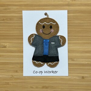 May include: A gingerbread person ornament with a grey jacket, blue shirt, and black trousers. The ornament has a white background and the text "Co-op Worker" is printed below the ornament.