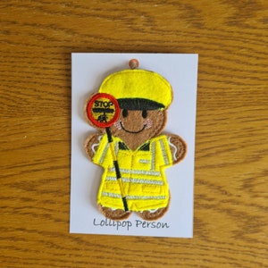 May include: A felt gingerbread person dressed as a lollipop person. The gingerbread person is wearing a yellow hat, a yellow vest with reflective stripes, and brown trousers. The gingerbread person is holding a red and white lollipop sign that says "STOP". The text "Lollipop Person" is written below the gingerbread person.