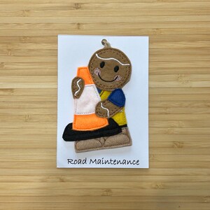 May include: A gingerbread person wearing a yellow vest and blue shirt holds a traffic cone. The text "Road Maintenance" is printed below the gingerbread person.