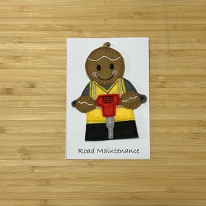 May include: A gingerbread person wearing a yellow safety vest and holding a red jackhammer. The gingerbread person has a brown face with a smile and brown eyes. The text "Road Maintenance" is written below the gingerbread person.