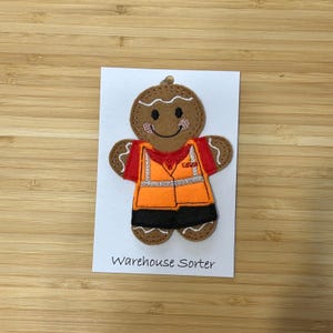 May include: A gingerbread person wearing a safety vest and black trousers. The gingerbread person is holding a sign that says "Warehouse Sorter".