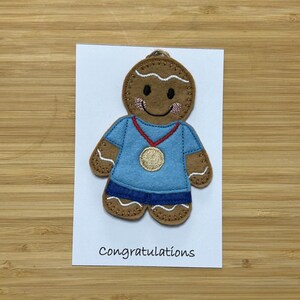 May include: A gingerbread person wearing a blue shirt and a gold medal. The gingerbread person is smiling and has red cheeks. The card says "congratulations".