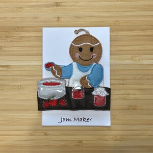 May include: A gingerbread person wearing a blue apron and holding a red strawberry. The gingerbread person is making jam and has three jars of jam on a table. The text "Jam Maker" is written below the image.
