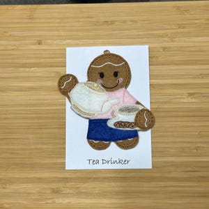 May include: A gingerbread person wearing a pink shirt and blue skirt is holding a teapot and a cup of tea. The gingerbread person is smiling and has brown eyes. The text "Tea Drinker" is below the gingerbread person.