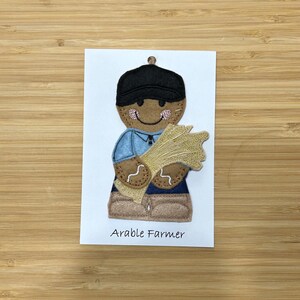 May include: A felt appliqué of a farmer holding a sheaf of wheat. The farmer is wearing a blue shirt, brown trousers, and a black hat. The text "Arable Farmer" is printed below the appliqué.