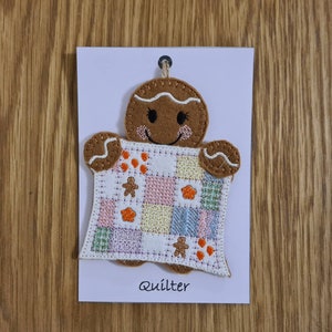 May include: A gingerbread person holding a patchwork quilt with a smiling face. The quilt is made of various colours and patterns. The gingerbread person is brown and has white icing. The text "Quilter" is printed below the gingerbread person.
