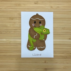 May include: A gingerbread man holding a green lizard. The gingerbread man is brown with white icing and has a smiling face. The text "Lizard" is written below the gingerbread man.