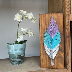 Könnte beinhalten: Eine Holztafel mit einem bunten String-Art-Federdesign. Die Feder besteht aus blauem, violettem und weißem Garn und ist vor einem braunen Holzhintergrund platziert.