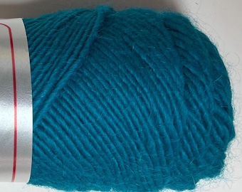 May include: A close-up of a ball of teal yarn. The yarn is tightly wound, showing the texture of the fibers. A white label with red stripes is visible on the side. This yarn is suitable for knitting and crochet projects.